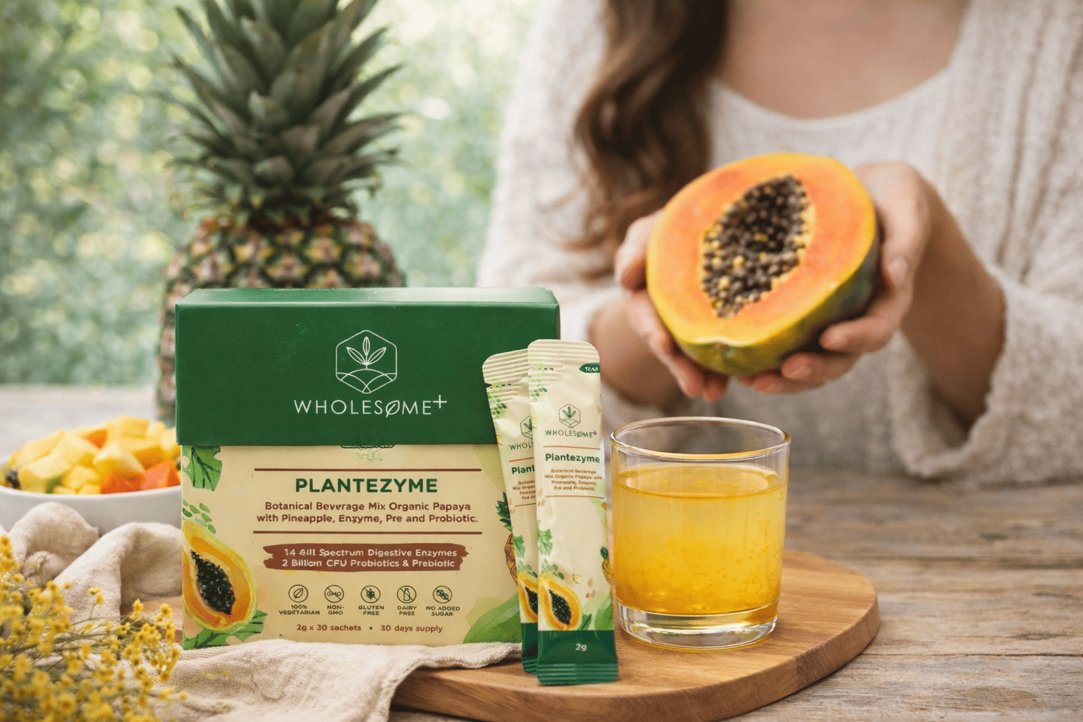 Wholesome+ Plantezyme box and sachets beside a glass of yellow drink, with a person holding a halved papaya and a pineapple in the background.